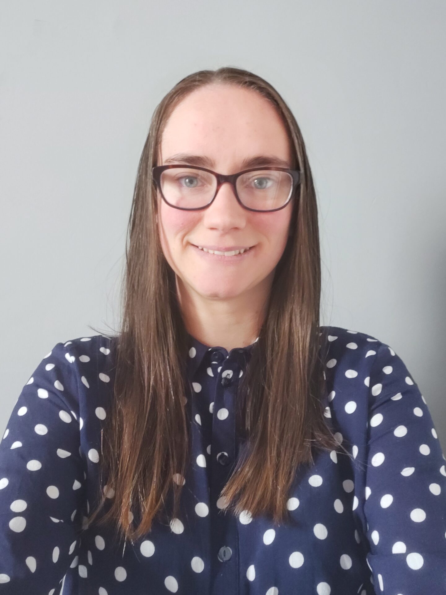 A woman with long brown hair and glasses is smiling at the camera, wearing a navy blue shirt with white polka dots, against a plain light gray background.