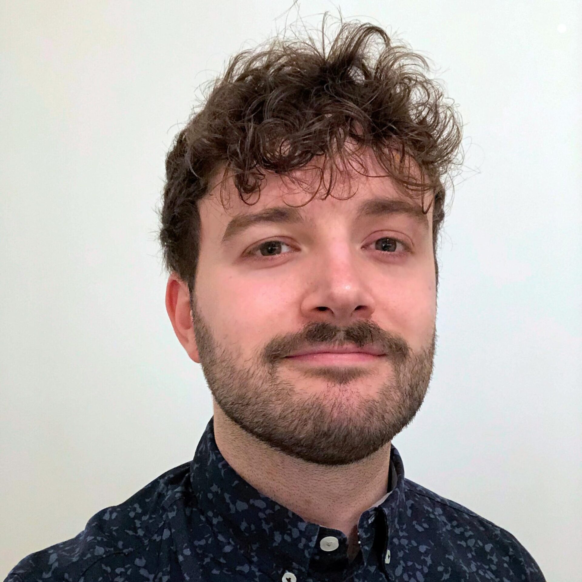 A man with curly brown hair and a beard is wearing a dark patterned button-up shirt and looking at the camera, standing against a plain light background.