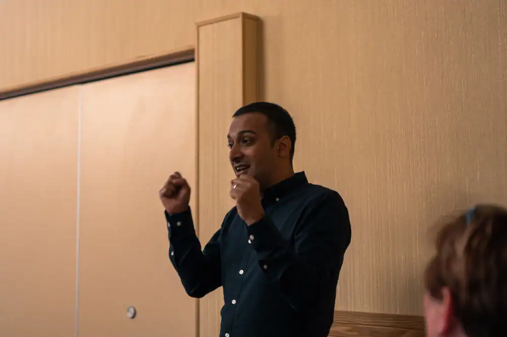 A person in a dark shirt stands indoors, gesturing with both hands raised, speaking to someone out of frame.