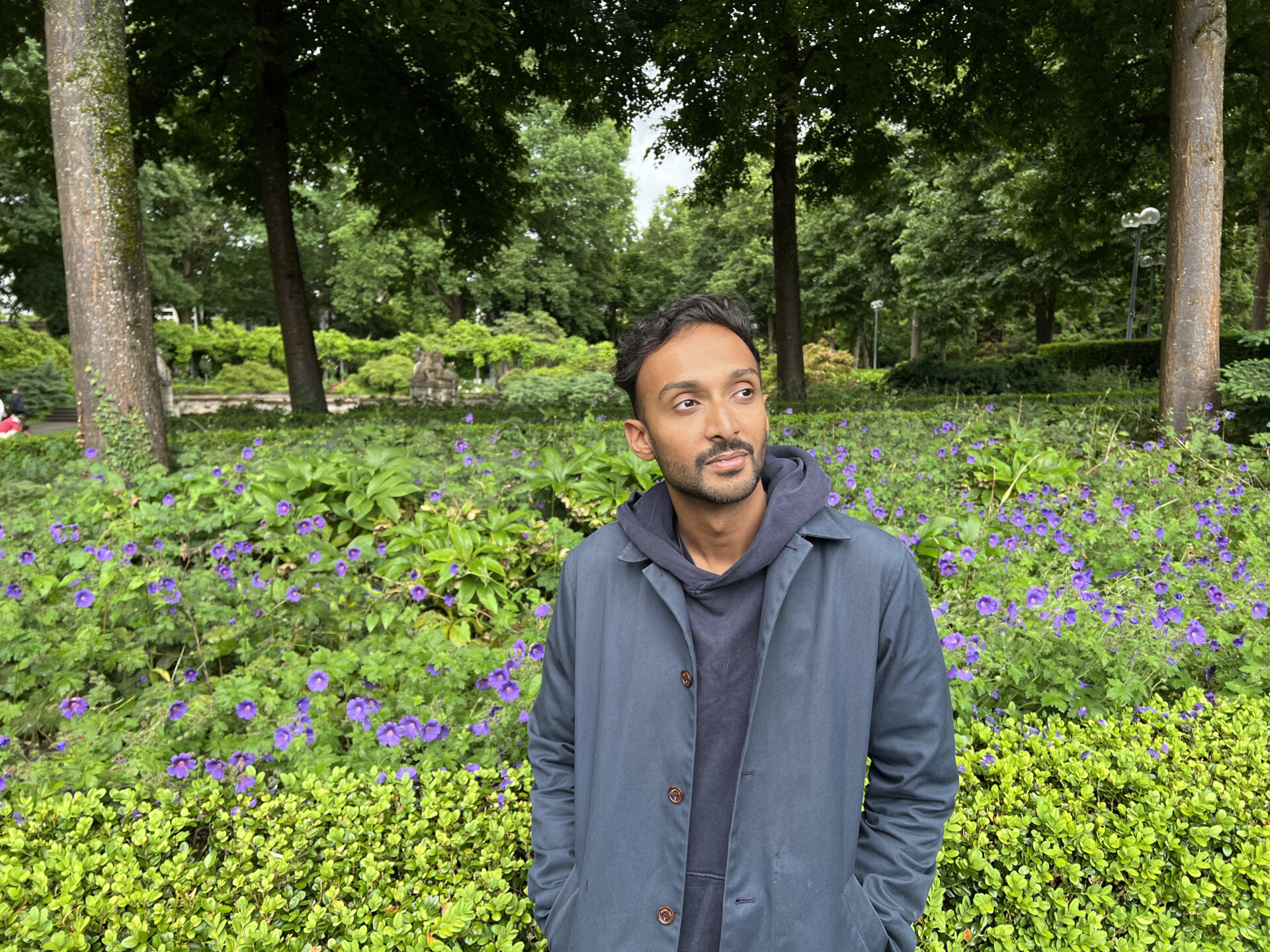 A man in a blue coat stands in front of a bush and purple flowers in a park, with tall trees and greenery in the background.