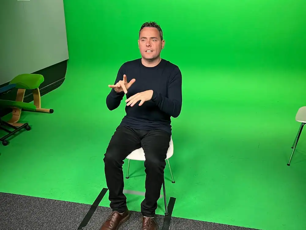 A person sits on a white chair in a studio with a green screen background, gesturing with their hands.