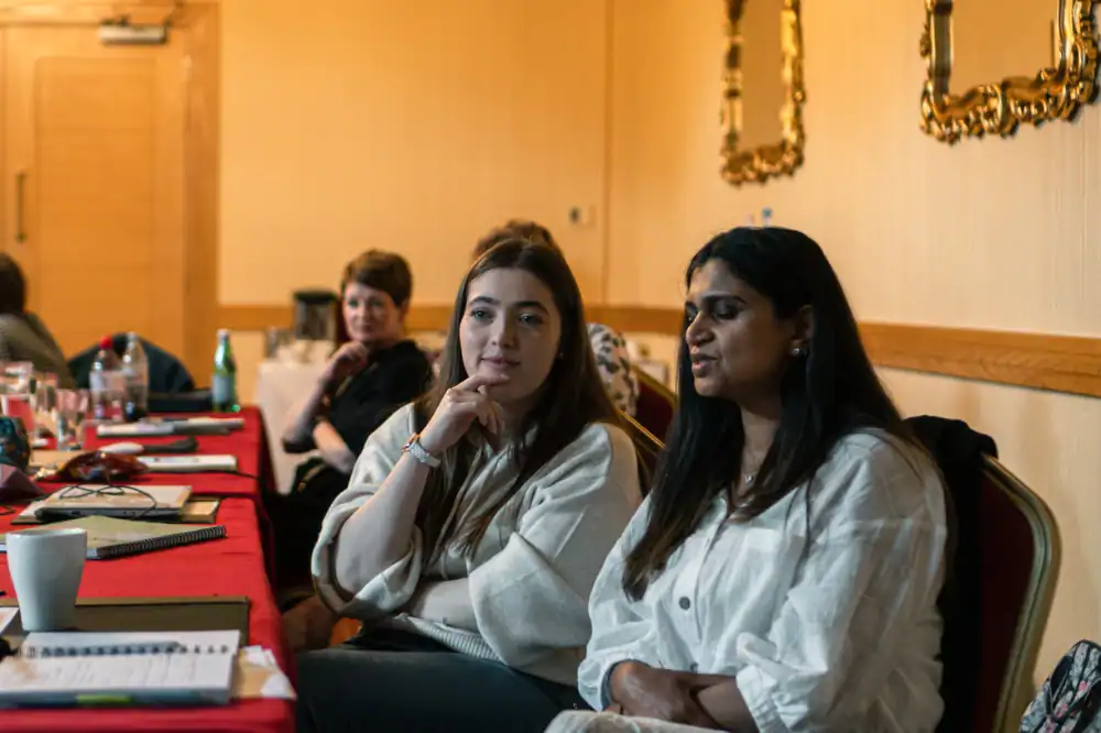 Several people sit at a long table in a warmly lit room, listening and conversing, with notebooks, cups, and writing materials on the table.