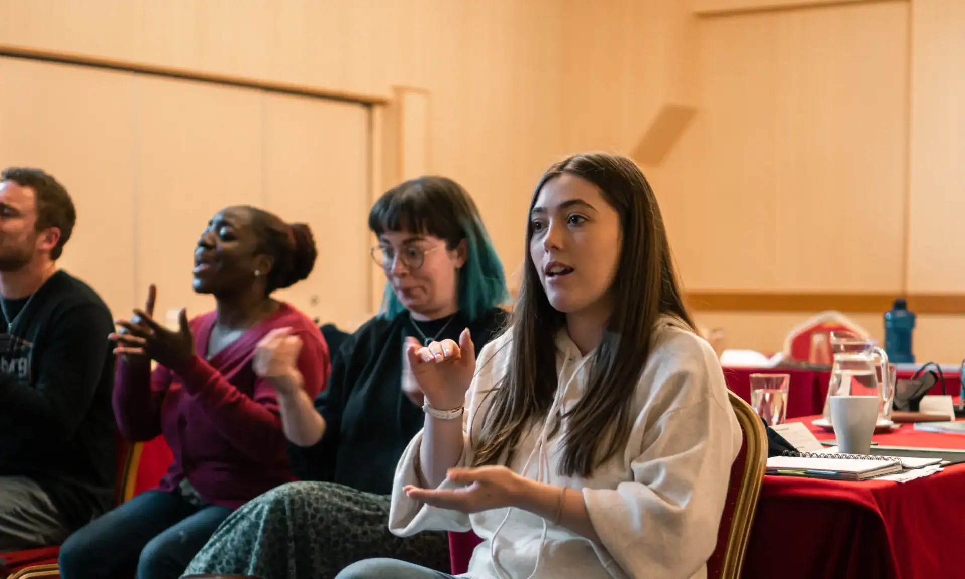 Several people sit in a room, actively using sign language and engaging in a discussion, with notebooks and cups on the table beside them.