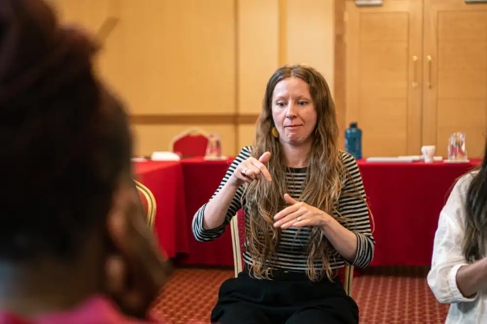 A woman with long hair and a striped shirt uses sign language while sitting in a conference room, facing two other people.