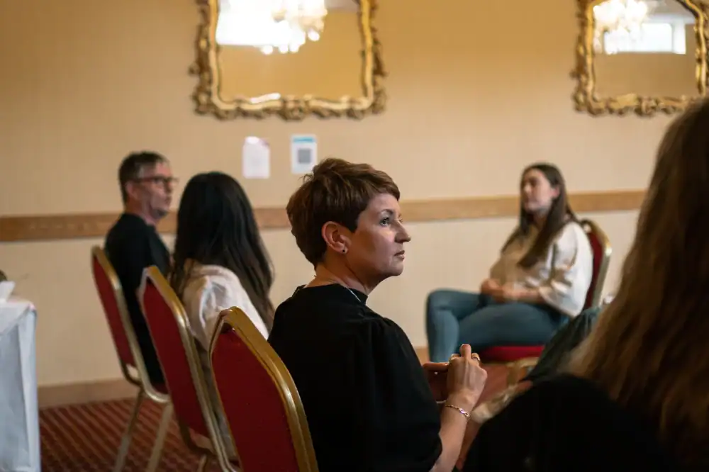 A group of people sit on red chairs in a room with ornate mirrors on the walls, engaged in conversation or listening attentively.