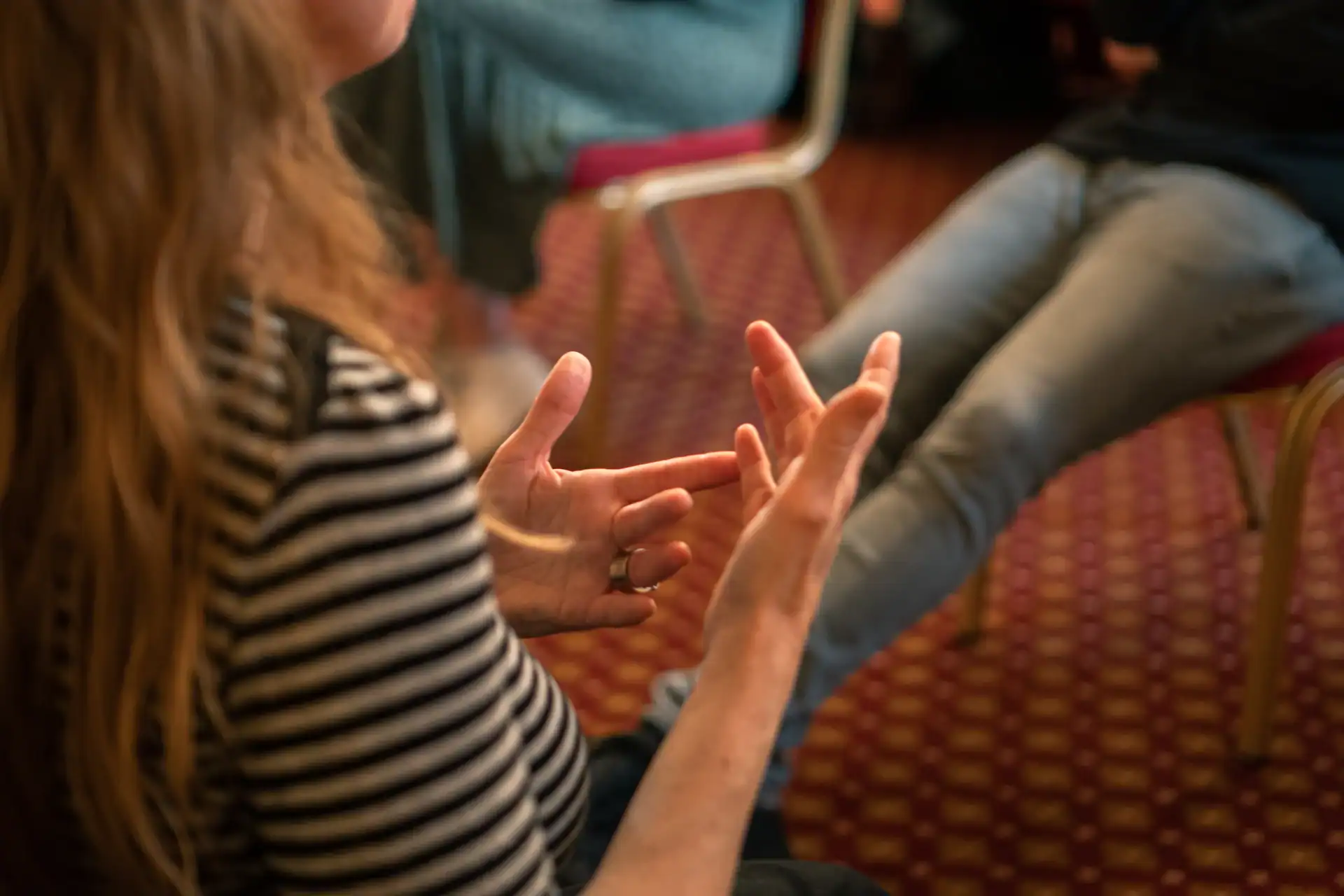 Person with long hair in a striped shirt gestures with their hands while talking to someone seated nearby on a red, patterned carpet.