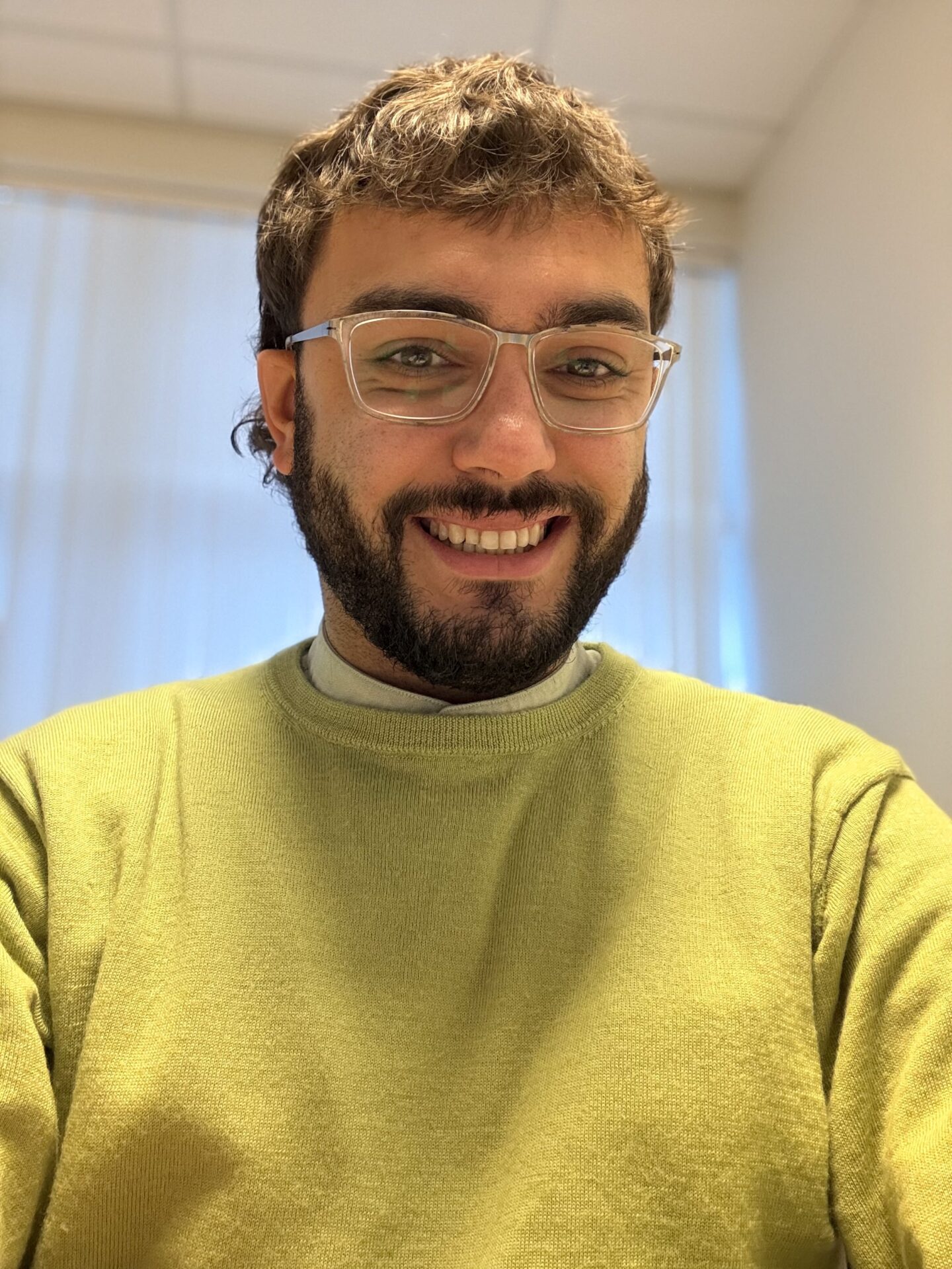 A person with short brown hair, glasses, and a beard is smiling at the camera, wearing a green sweater in a brightly lit room.