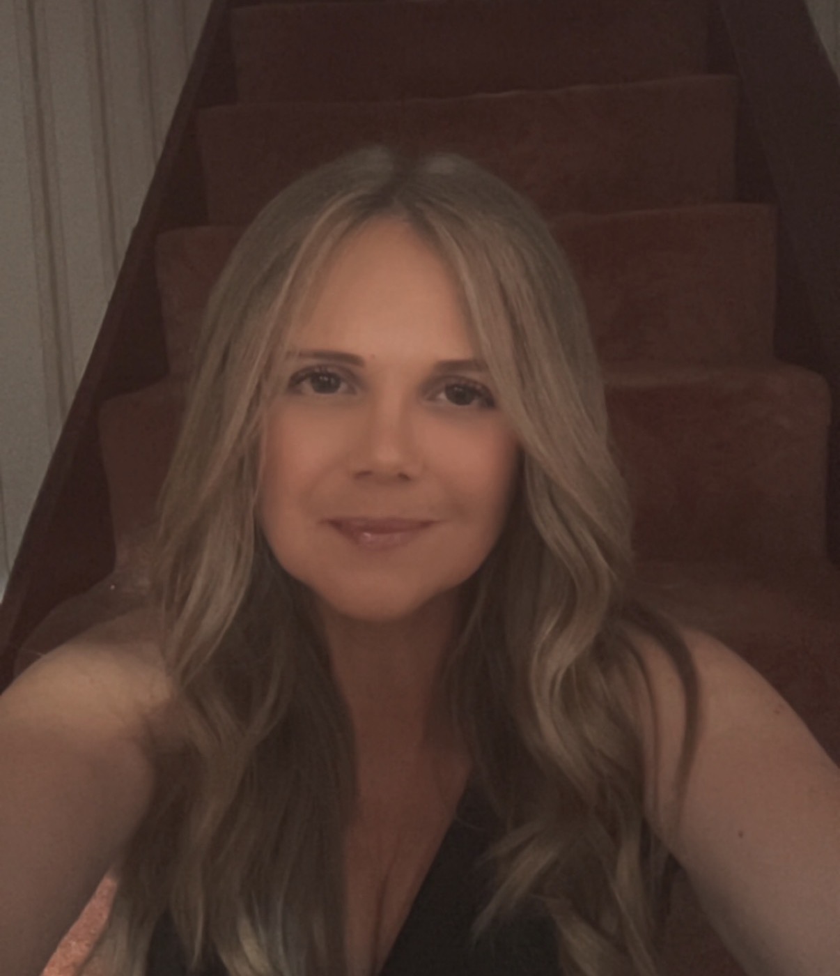 A woman with long blonde hair is sitting on carpeted stairs, looking at the camera and smiling slightly.