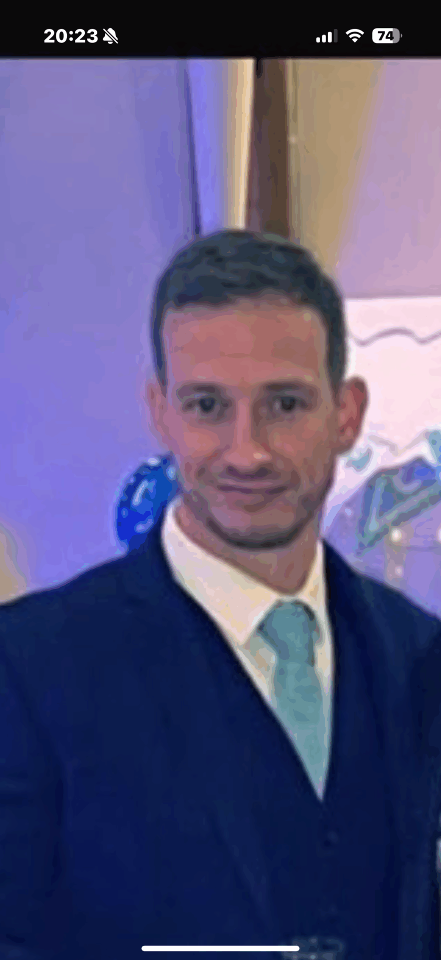 A man in a suit and light blue tie is standing indoors, facing the camera. The image has a filtered, painted effect.