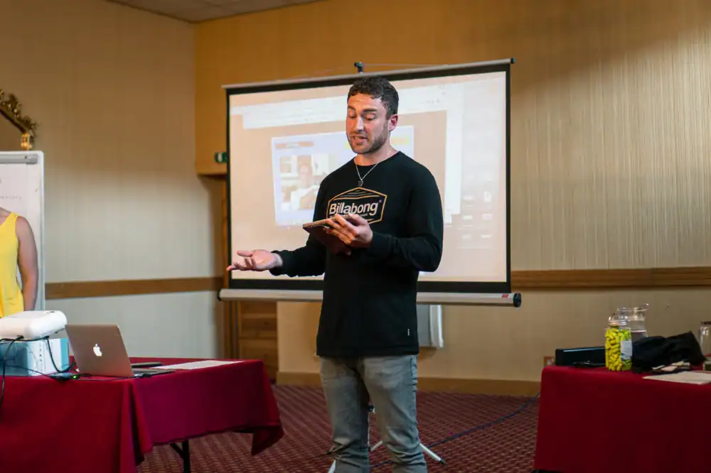 A man in casual clothing gives a presentation using a tablet, standing in front of a projection screen in a conference room.