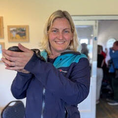 A woman in a blue jacket smiles at the camera while holding a small, dark object in her hands indoors.