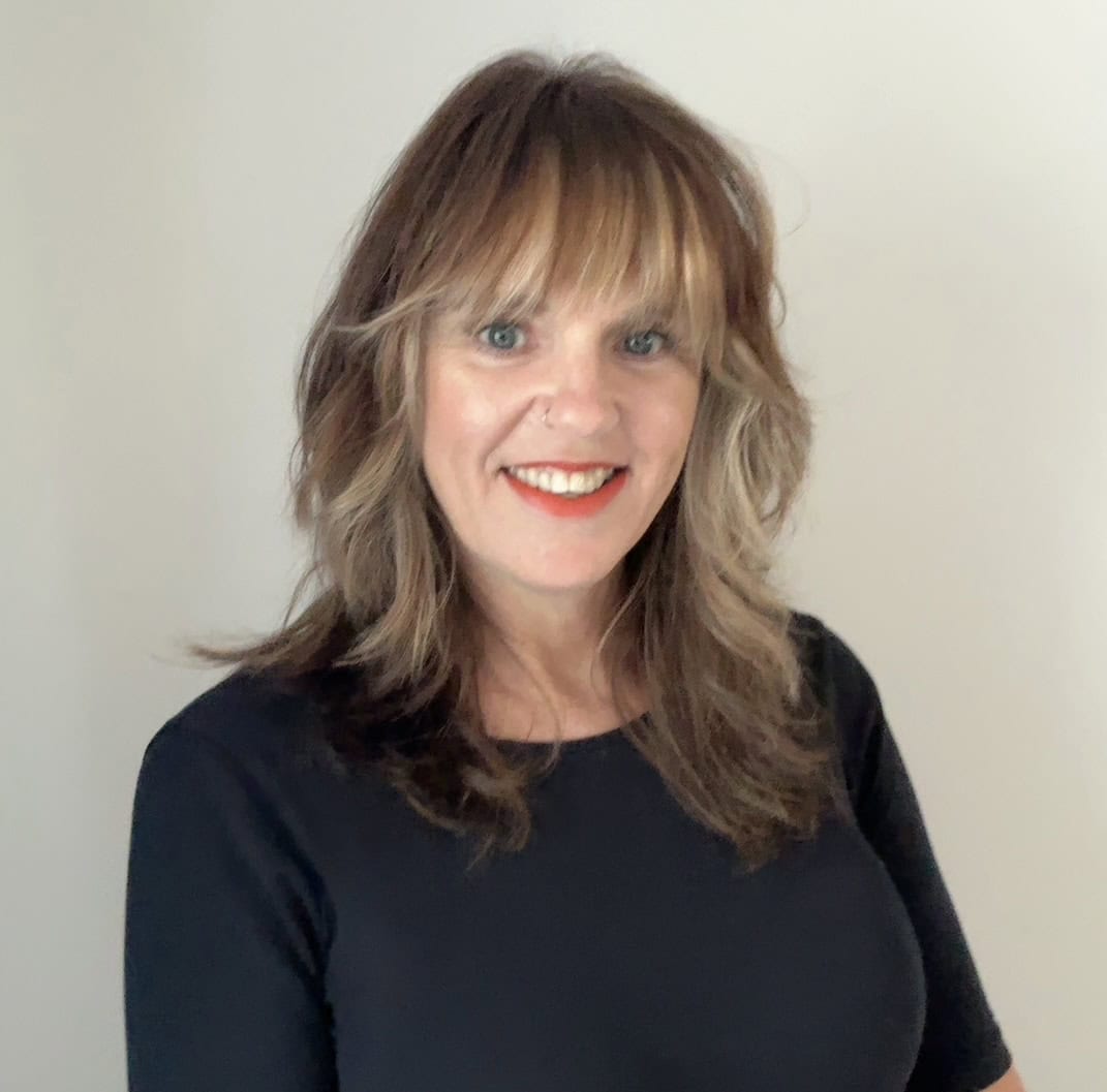 Woman with shoulder-length brown hair and bangs, wearing a black top, standing in front of a plain light-colored background and smiling.