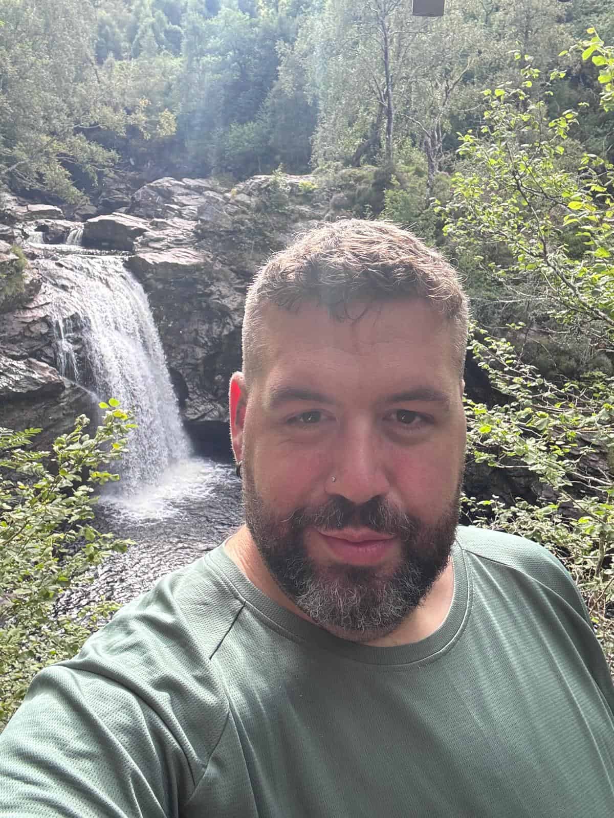 A man with a beard and short hair takes a selfie outdoors with a waterfall and lush greenery in the background.