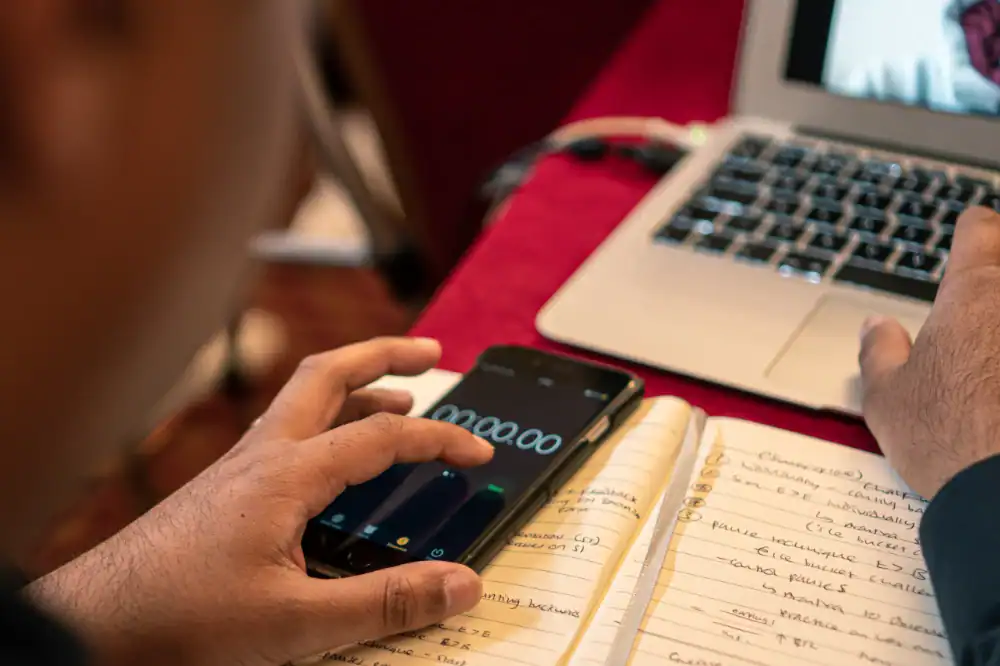 A person uses a smartphone stopwatch while taking handwritten notes beside an open laptop on a red table.