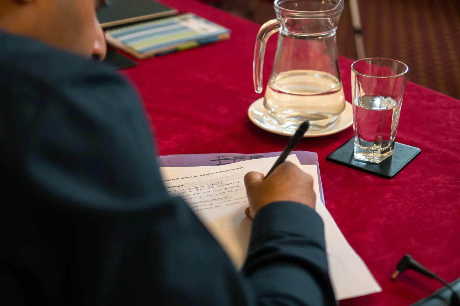 Person writing on a document at a table with a glass and a pitcher of water, notebooks, and a pen on a red tablecloth.