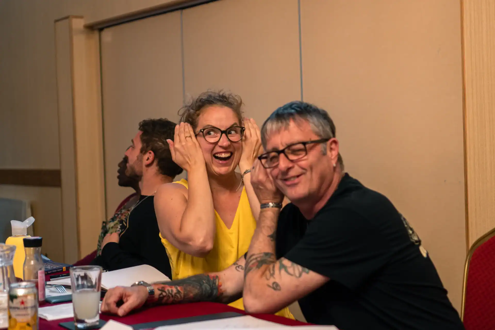 Two people sit at a table smiling and holding their hands up to their heads, while another person sits behind them looking away. Papers and drinks are on the table.