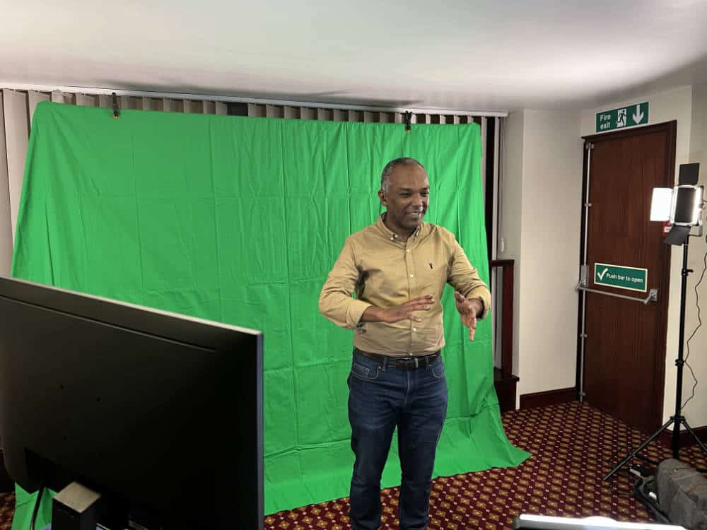 A man stands in front of a green screen in a room with studio lights, a monitor, and patterned carpet, appearing to speak or present.