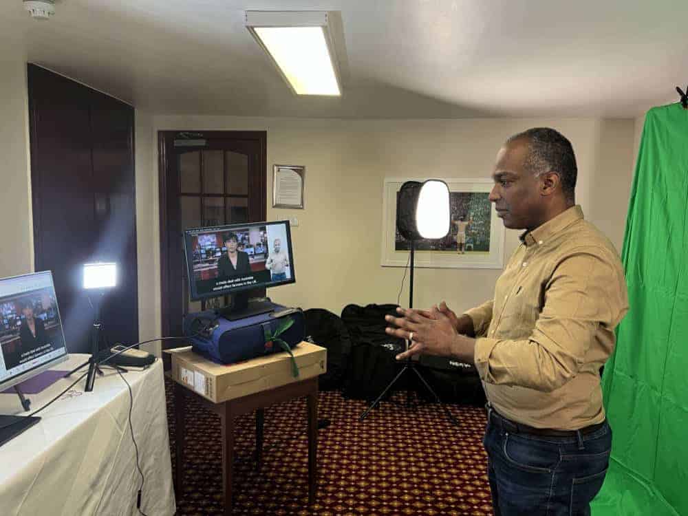 A man stands in a small room with video equipment, speaking in front of a computer and camera, with lights and a green screen set up behind him.