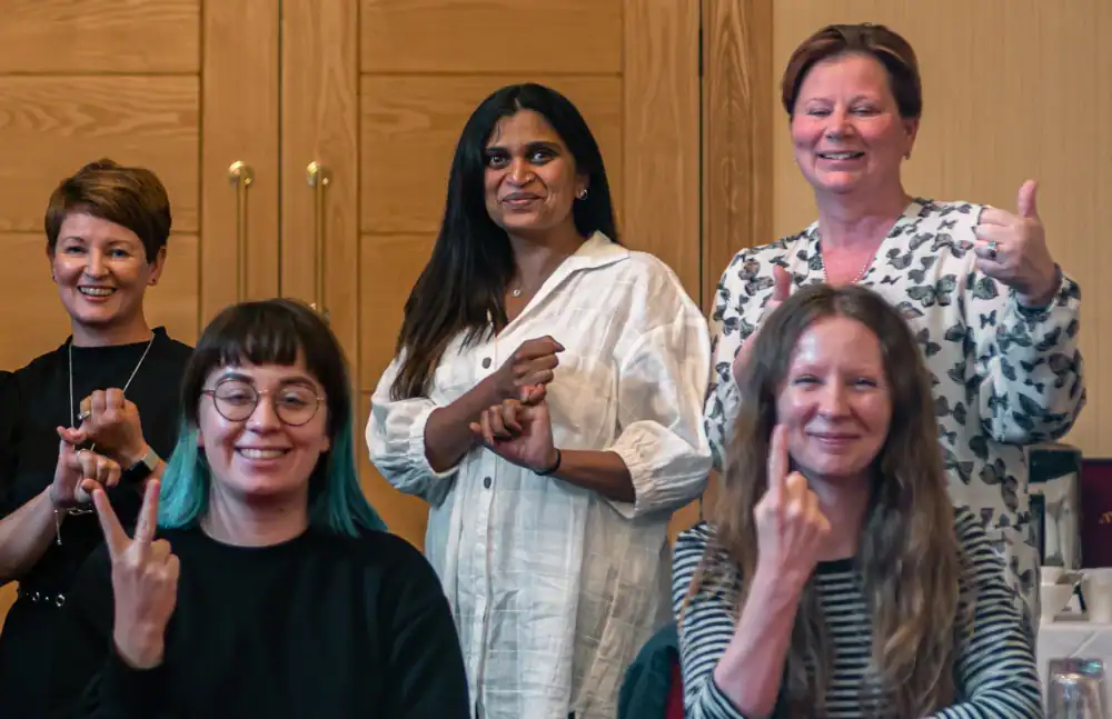 Five people, including four women standing and one sitting, pose indoors, with three making sign language gestures and two smiling at the camera.